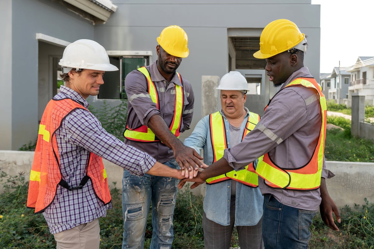 Four construction workers joining hands on site, wearing hardhats and safety vests